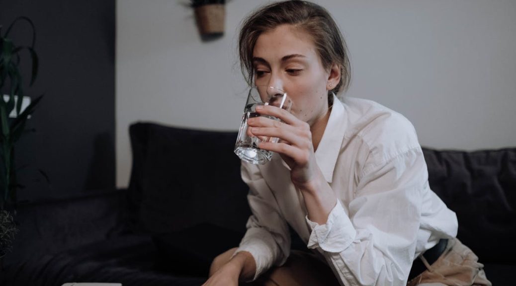 Woman in a white shirt sitting on a sofa, drinking water at home for relaxation.